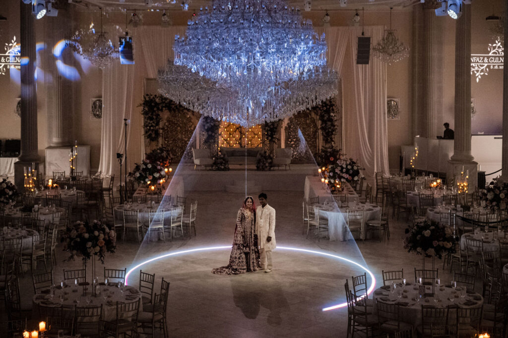 Bride and groom stand for posed portrait in the middle of the grand ballroom of the bellevue surrounded by lasers and chandeliers